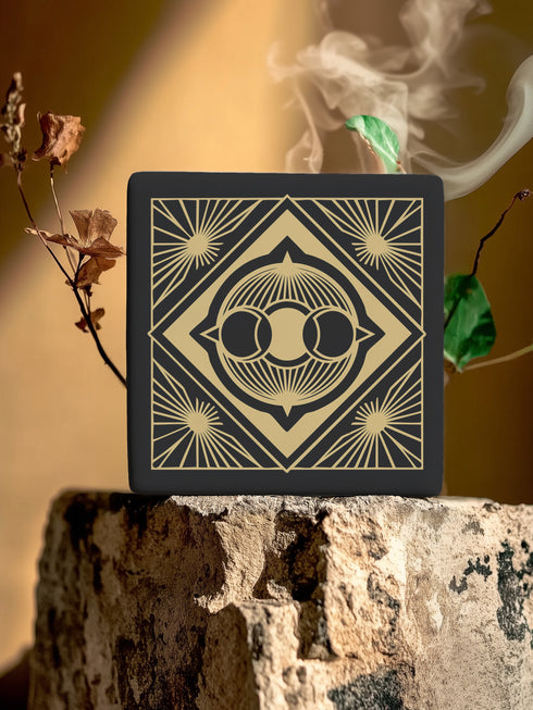 A black and gold ceramic coaster featuring a triple moon design framed by art deco geometric details and celestial sunburst accents.